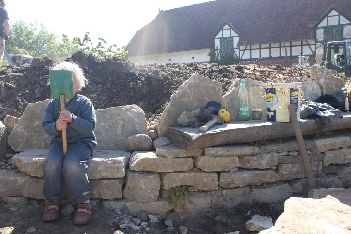 Eine Bank gebaut aus Bruchstücken der Betonbodenplatte, darauf Gehörschutz, wie ihn Bauarbeitende tragen sowie zwei Falschen. Am linken Bildrand sitzt ein Kind, das sich hinter einer Schaufel versteckt.