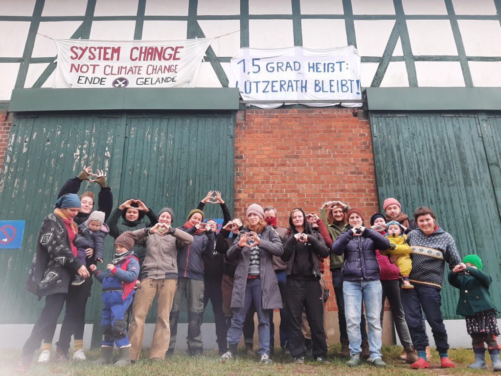 Bewohner*innen des Raeume Wohnprojekts stehen vor grünen Scheunentoren. Mit ihren Händen formen sie Herzen. Über ihren Köpfen hängen zwei Banner. "System change, not climate change" steht auf dem einen, auf dem anderen steht "1,5 Grad heißt: Lützerath bleibt!"
