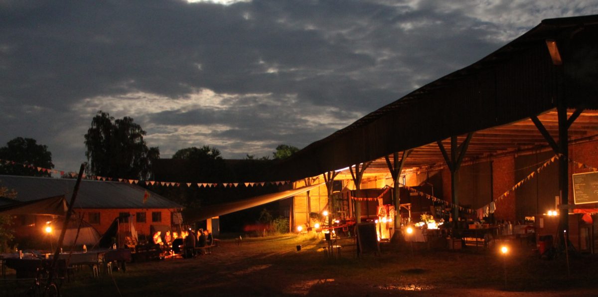 Vor einem dunkler werdenden Sommerhimmel steht ein erleuchtetes Scheunengebäude. Eine Wimpelgirlande verbindet das Gebäude mit etwas links außerhalb des Bildausschnitts.