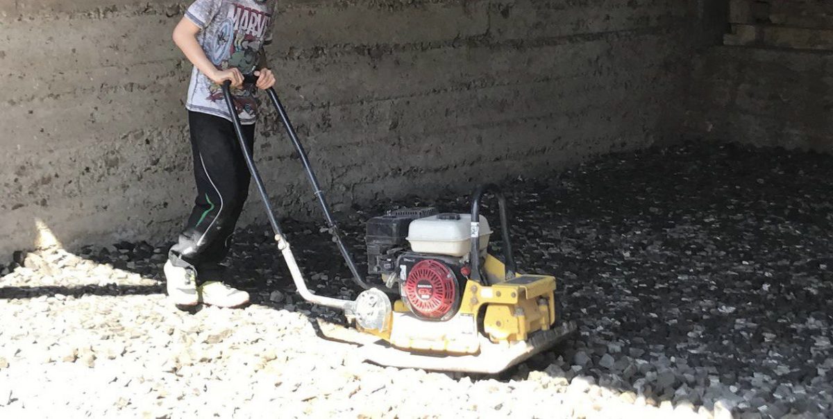 Eine Person, auf dem Bild ohne Kopf, schiebt eine Rüttelmaschine über eine Glasschaumschotterfläche.