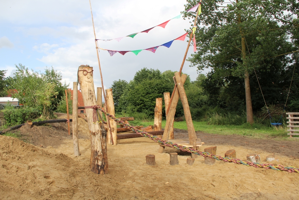 Nebn einem großen Sandkasten stehen mehrere Spielgeräte aus Holzstämmen.